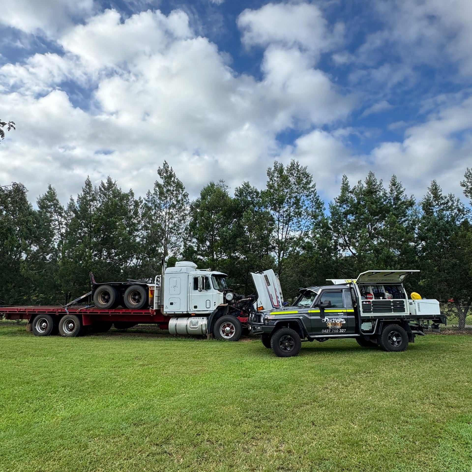Truck with a trailer carrying large equipment on a grassy field with trees and blue sky. Being worked on by a ute with tool boxes